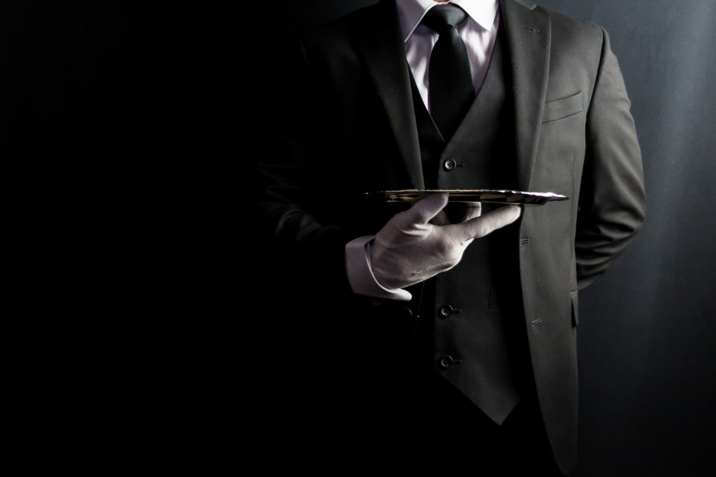 Portrait of a formal butler in a dark suit and white gloves holding a silver serving tray, representing professional hospitality and courtesy.